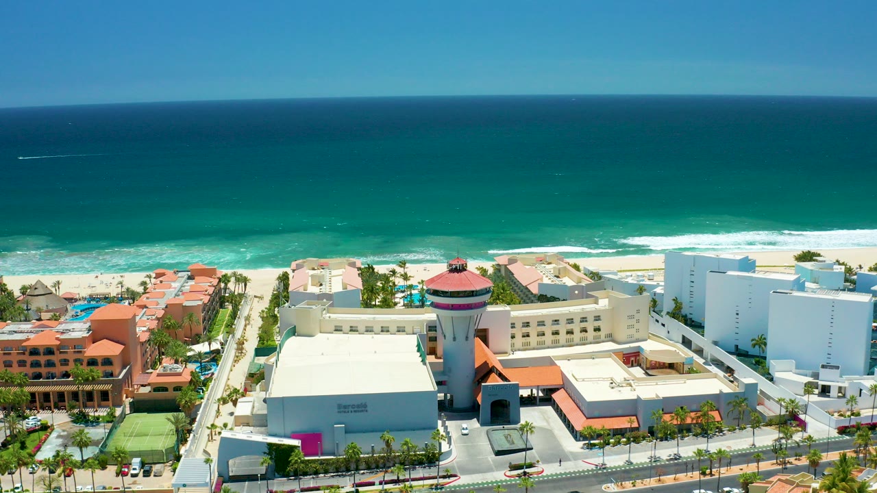 High aerial frame over San Jose Del Cabo resort buildings, pools, palm trees, sandy beach, and turquoise ocean with no visible people.