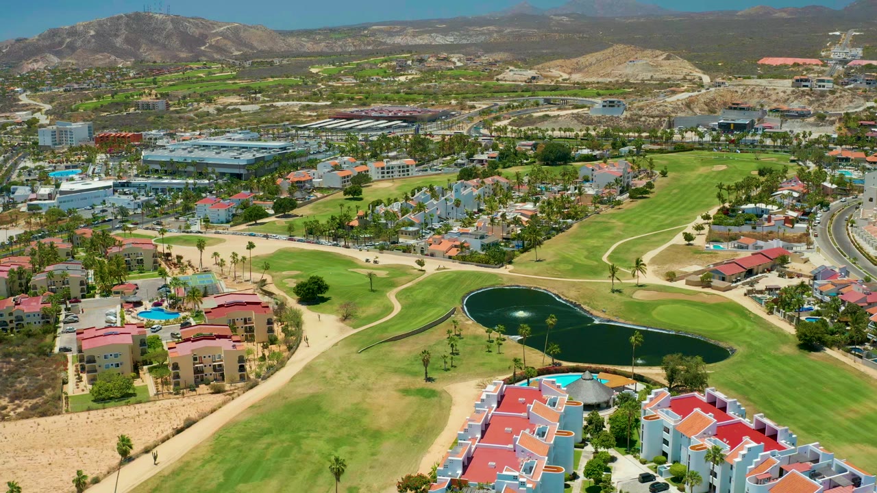 Early aerial frame over a San Jose Del Cabo golf course with a dark pond near the lower center, pale fairway grass, red-roofed buildings in front, distant hills, and no visible people.