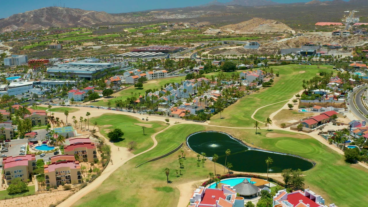Early aerial frame over San Jose Del Cabo showing a dark pond below green fairways, residential buildings, roads, desert hills, distant mountains, and no visible people.
