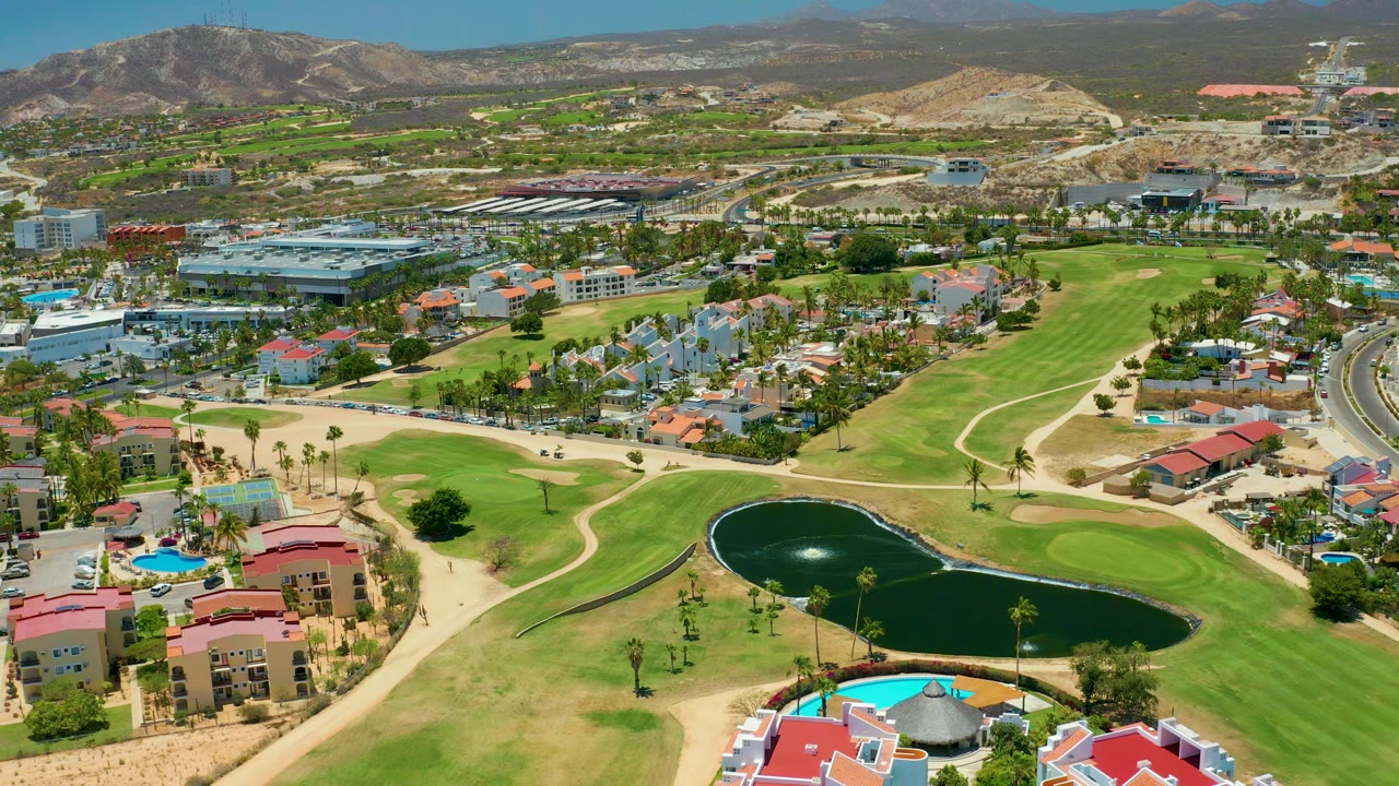 Aerial view in San Jose Del Cabo with the pond near the lower center, curved golf paths, palm-lined housing, pools, roads, dry hills, and no visible people.