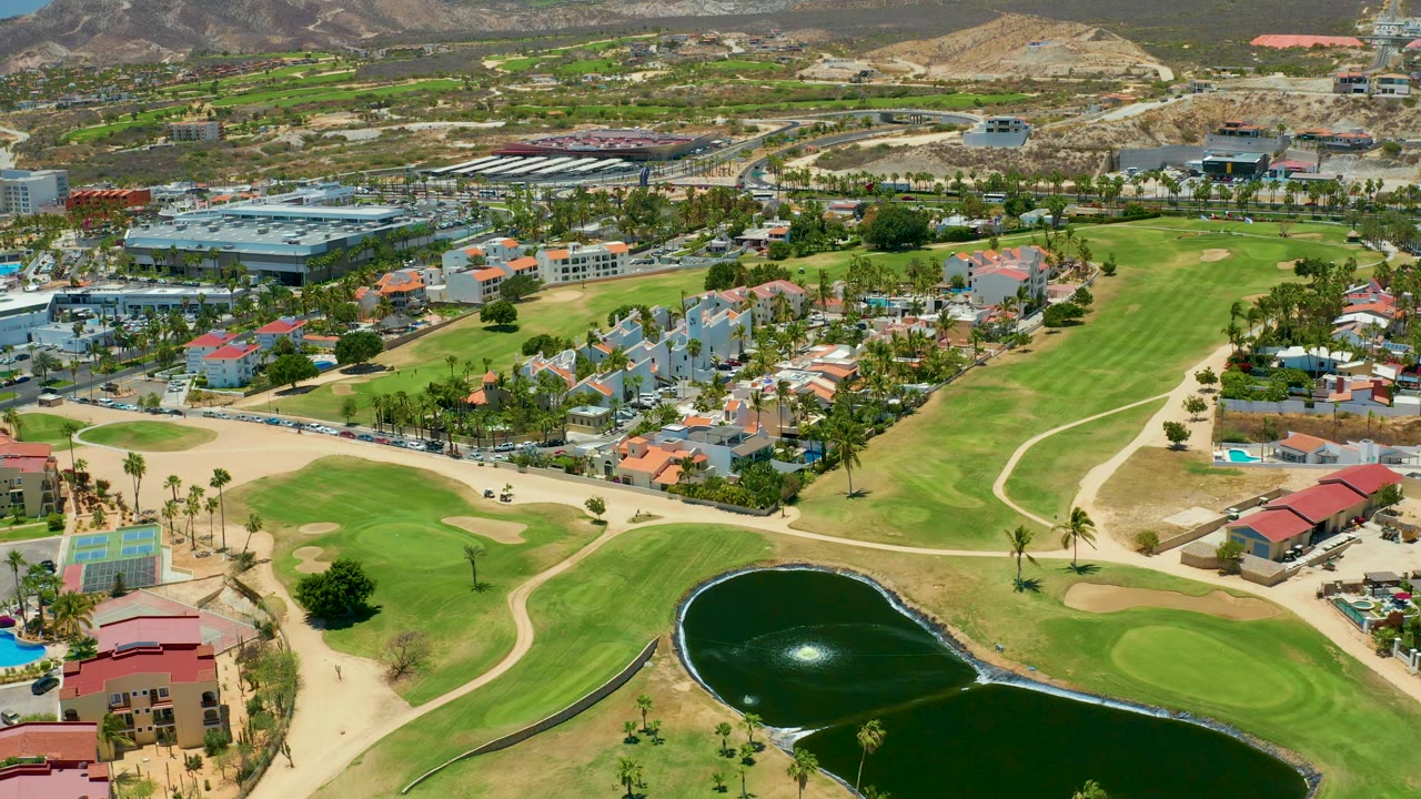 Aerial frame in San Jose Del Cabo showing the pond slightly more centered below the fairway, with residential buildings, palm-lined streets, parked vehicles, and no visible people.