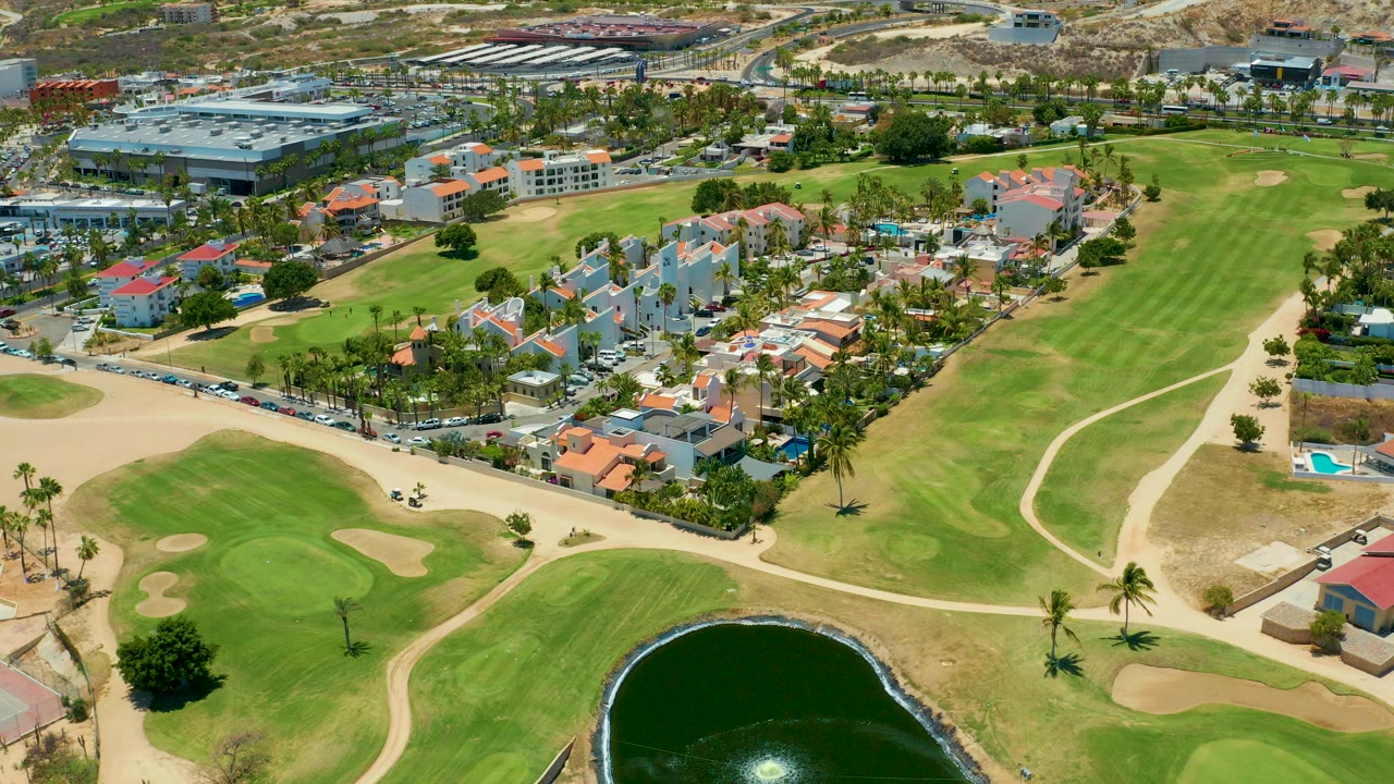 At 0:00, an aerial view in San Jose Del Cabo shows green golf fairways, sand traps, a dark pond edge, roads, palm trees, and red-roofed homes with no visible people.