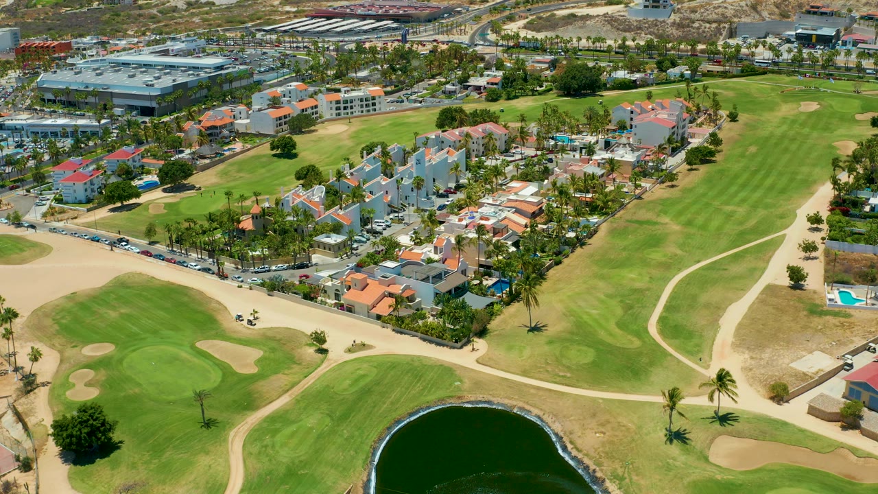 At 0:02, the aerial frame shows the golf course pond lower in view, wide fairways and sand traps across the center, and San Jose Del Cabo residential blocks and roads beyond with no visible people.