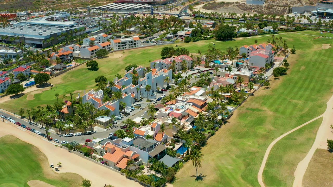 Aerial view in San Jose Del Cabo, Baja California Sur showing red-roof homes, palms, parked cars, sand areas, green fairway, and no visible people.