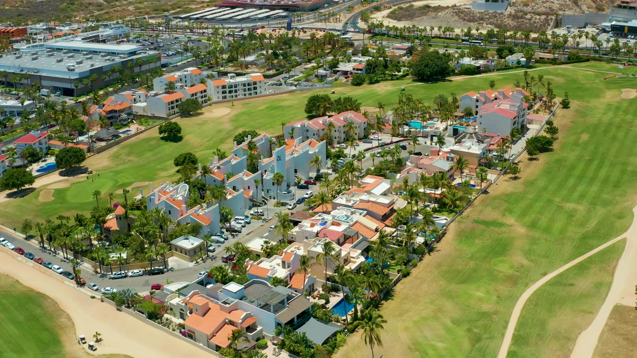 Aerial view in San Jose Del Cabo with clustered homes, visible pools, palm-lined streets, golf greens, sand traps, and no visible people.