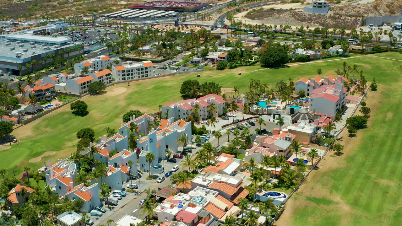 Aerial frame over San Jose del Cabo showing white residential buildings with red-tiled roofs, green golf course grass, palms, parking areas, and no visible people.