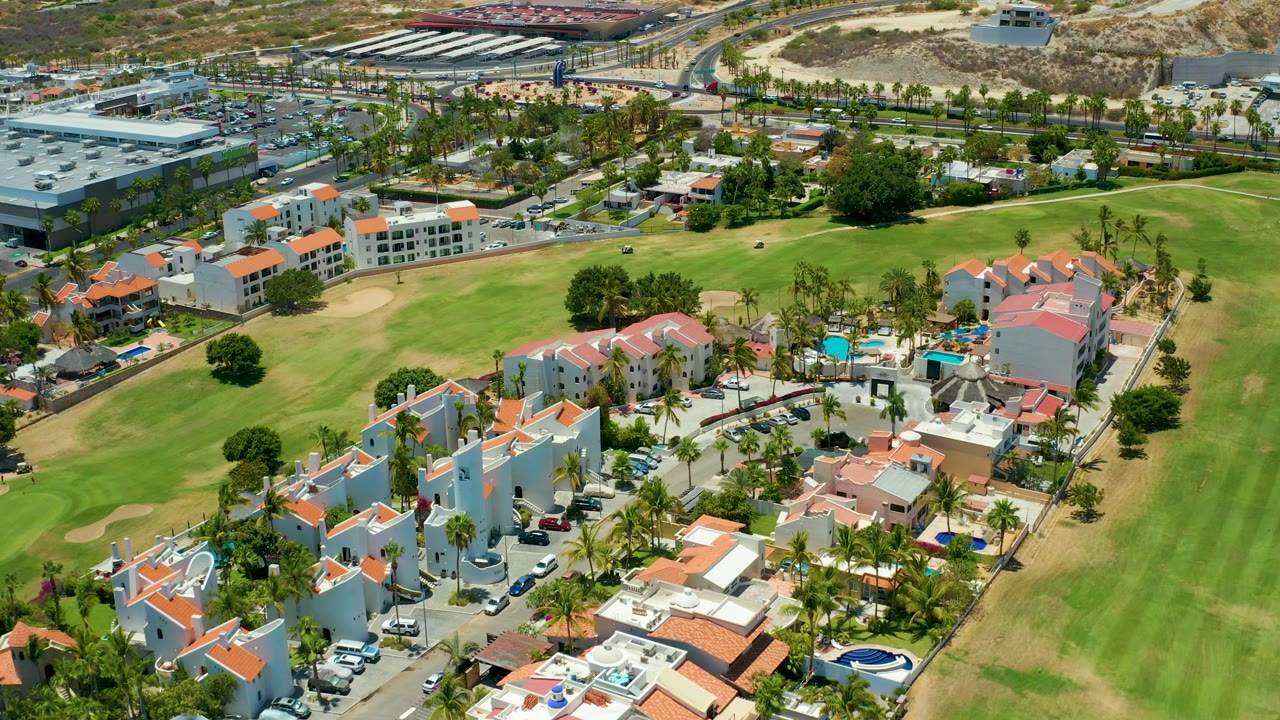 San Jose del Cabo aerial still with white walls, red-tiled roofs, a broad green golf course area, surrounding streets, commercial parking, dry hillside terrain, and no visible people.
