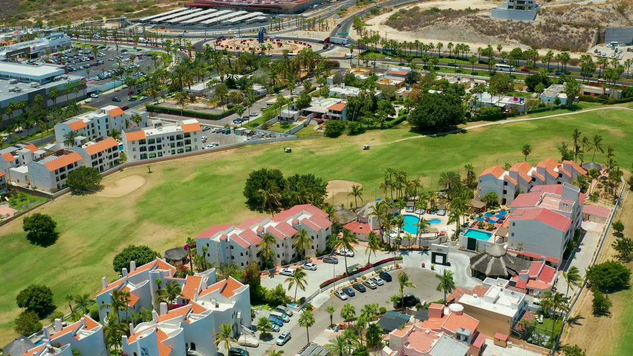 Aerial view over San Jose Del Cabo resort roofs and pool deck, with the golf course filling the middle of the frame, a parking area beyond it, and no visible people.
