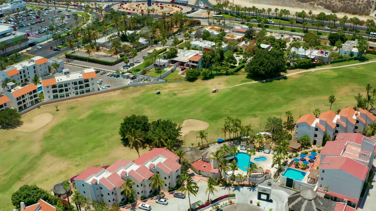 Aerial opening frame over a San Jose Del Cabo resort golf course with green fairway, tan dry patches, sand traps, red-roofed buildings, pools, palms, roads, cars, and no visible people.