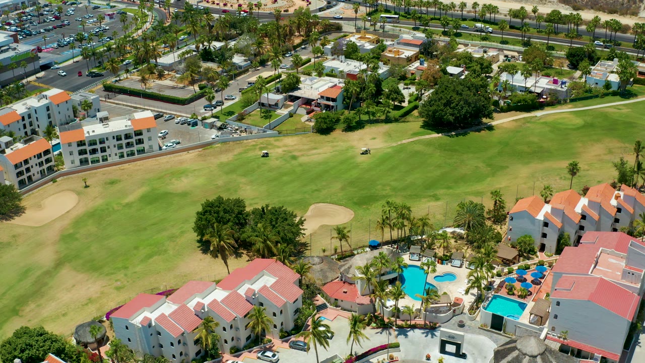 Aerial frame over San Jose Del Cabo with a broad green fairway, smaller brown turf areas, white resort buildings with orange-red roofs, two bright pools, palms, roadside homes, and no visible people.