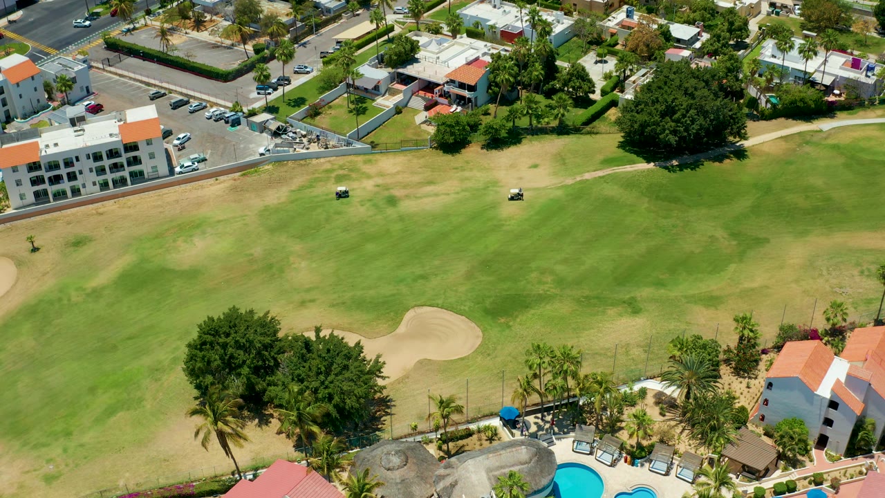 Aerial frame in San Jose del Cabo looking across a broad golf fairway where two carts sit against green grass, a sand trap, roadside buildings, and parked cars, with no visible people.