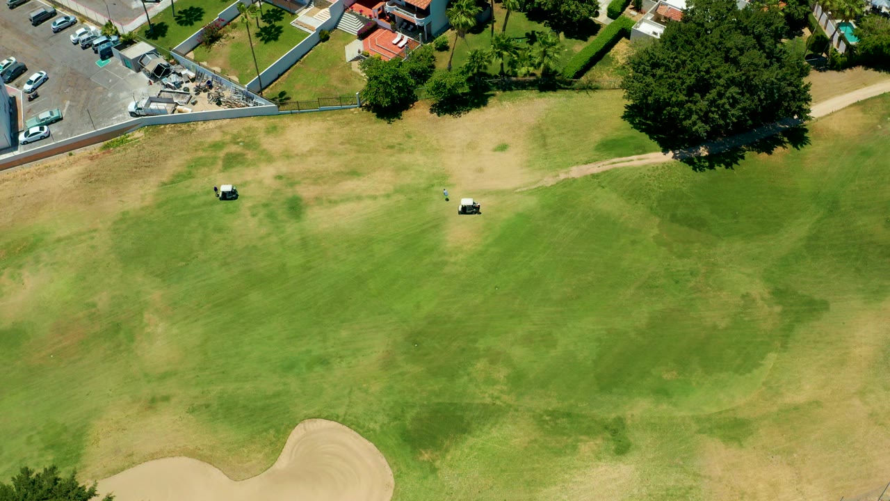 Opening aerial frame in San Jose del Cabo showing a green and tan golf fairway, two small golf carts, a curved sand bunker at the lower edge, parking and trees above, and no visible people.
