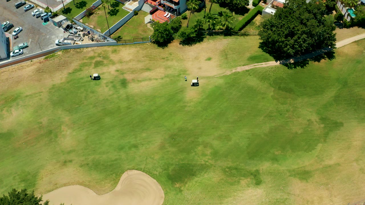 Overhead view in San Jose Del Cabo, Baja California Sur, showing a patchy green golf fairway with two golf carts, a sand trap at the bottom edge, parking lot cars, trees, buildings, and no visible people.