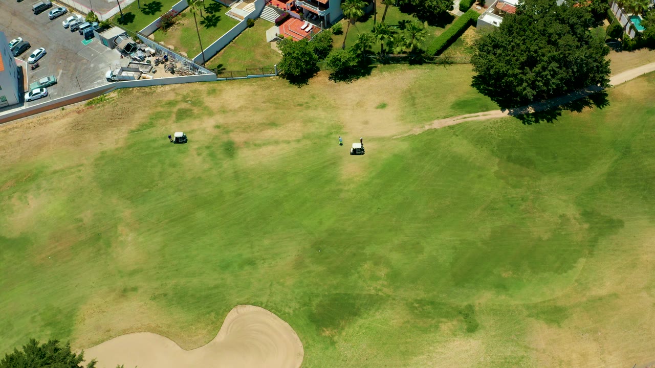 Aerial fairway scene with golf carts, patchy grass, sand trap, parking lot, trees, and buildings.
