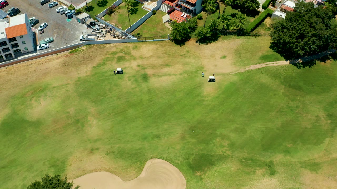 A high aerial frame in San Jose del Cabo shows two white golf carts separated across the fairway, patchy grass below, a parking area and low buildings above, and no visible people.