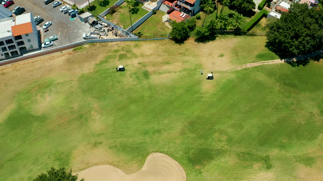 San Jose del Cabo aerial frame with two golf carts on a mottled green and brown golf course, trees and buildings along the top boundary, parked cars nearby, and no visible people.