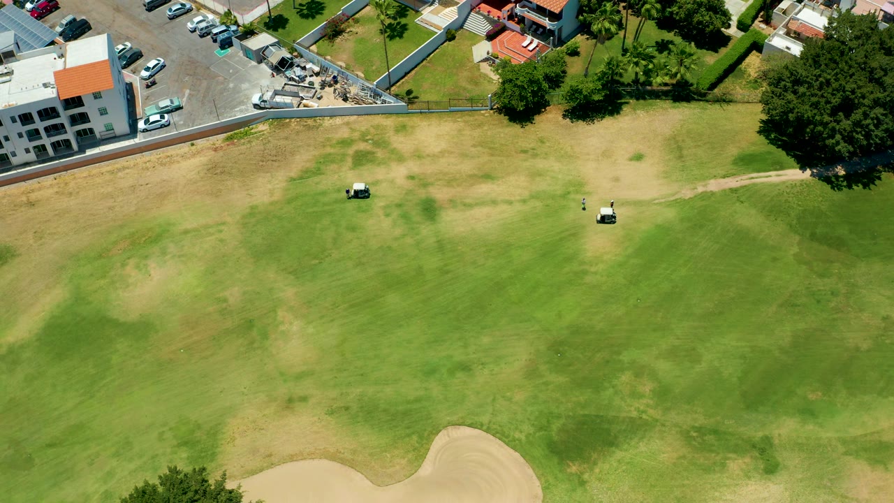 Golf carts crossing a dry fairway area near a parking lot and white building.