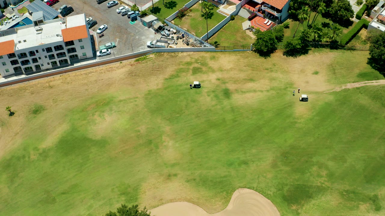 Overhead view in San Jose Del Cabo, Mexico, showing two golf carts on a patchy green and brown fairway, a sand trap at the lower edge, and buildings with parked cars along the top, with no visible people.