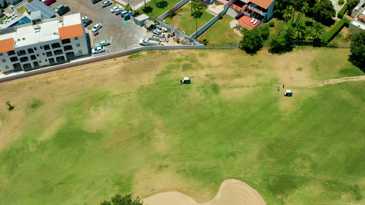 Aerial frame from San Jose Del Cabo, Mexico, with the golf carts slightly farther across the fairway, dry brown patches in the grass, and the parking lot and buildings still visible at the top, with no visible people.