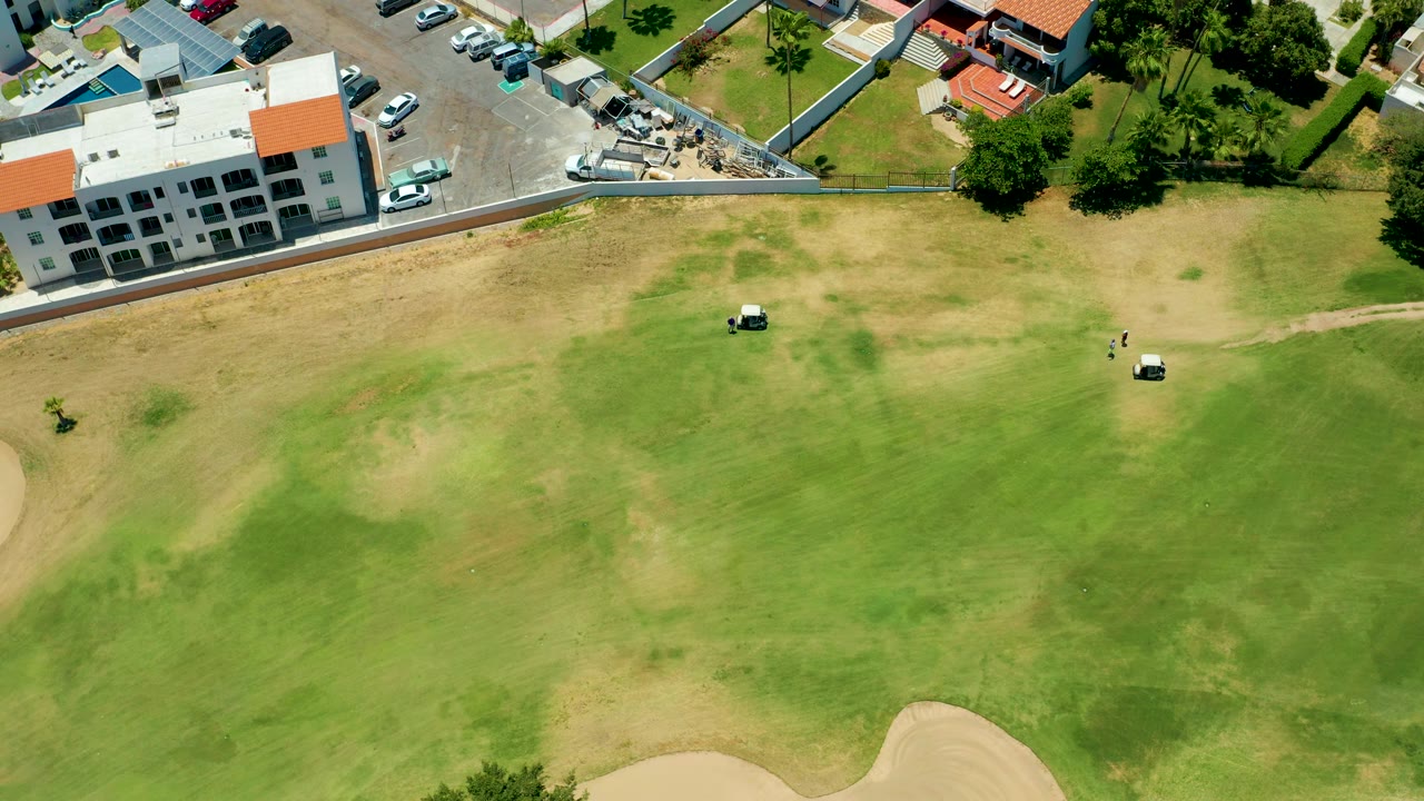 Drone view in San Jose Del Cabo, Mexico, with two golf carts separated on the fairway, a pale sand trap near the lower right, and buildings, trees, and parked vehicles along the upper border, with no visible people.