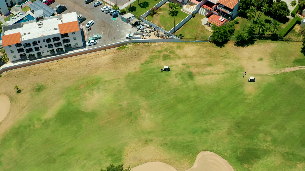 Wide aerial view of golf carts on patchy course grass beside a parking lot.