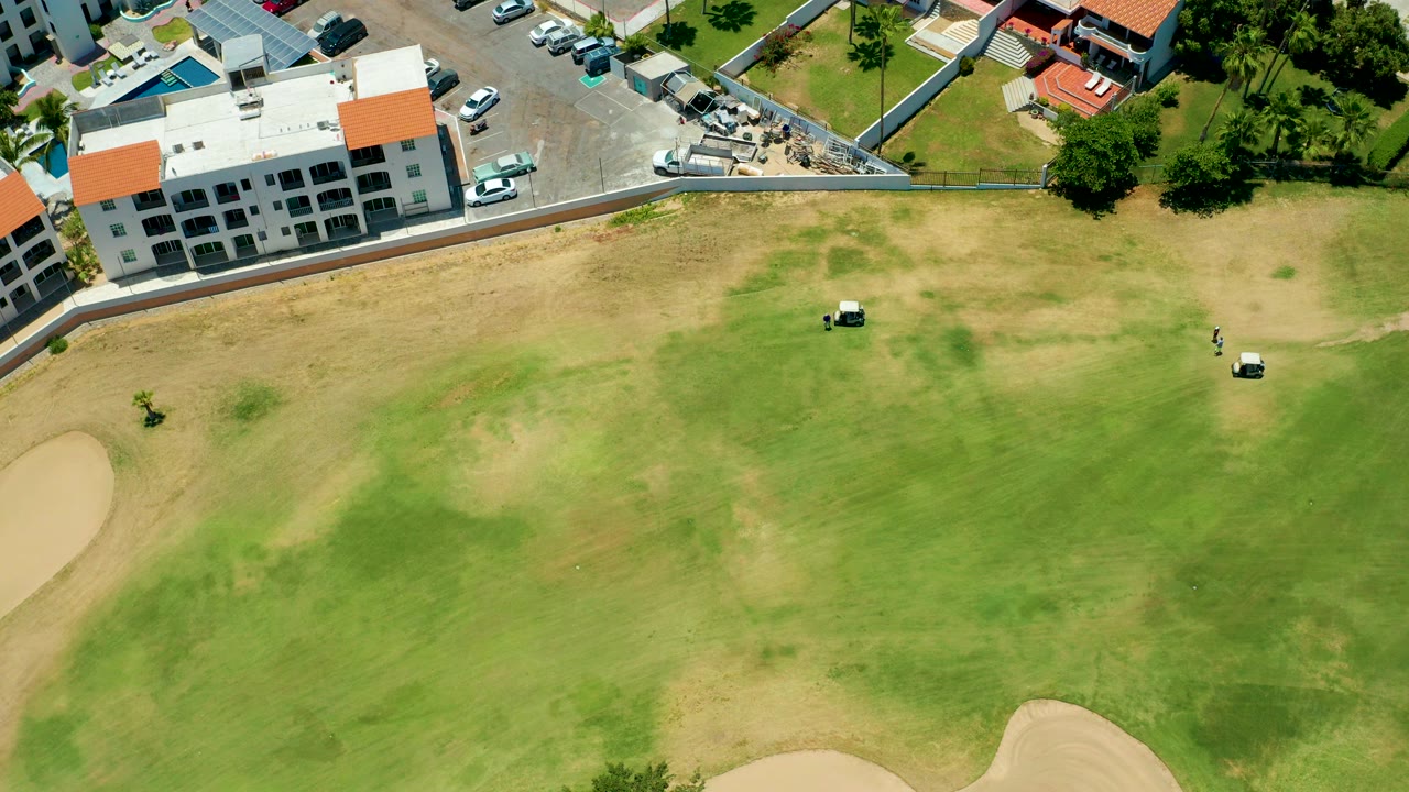 Overhead view of golf carts on a patchy green beside buildings and sand traps.