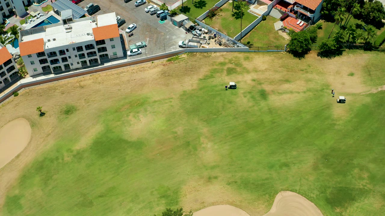 Aerial frame from San Jose del Cabo showing two small golf carts farther across the green, tan bunkers at the lower edges, parked vehicles by a white building, and no visible people.