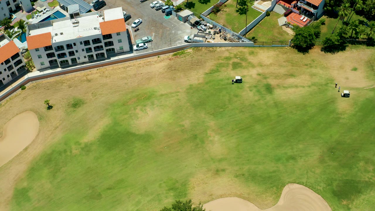 Overhead San Jose del Cabo frame with one golf cart near the center of the patchy green, another closer to the right edge, a sand trap at lower left, buildings above, and no visible people.
