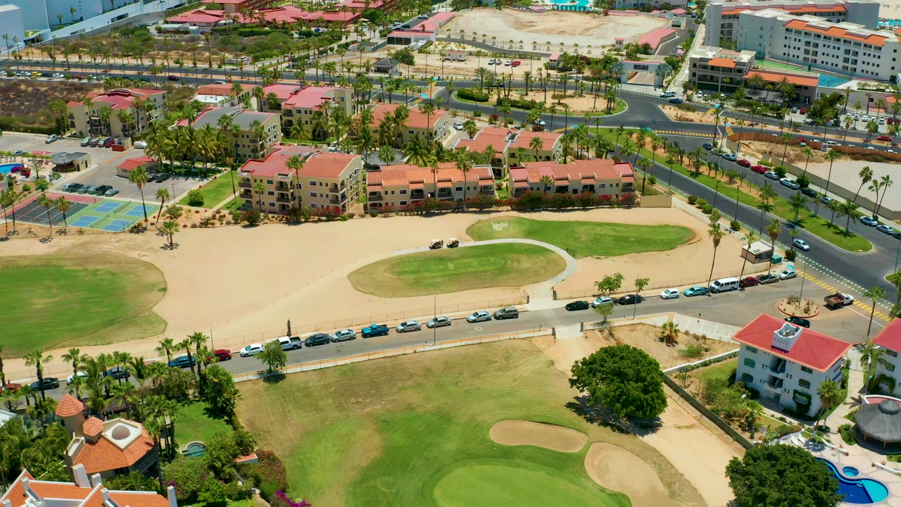 Opening aerial frame over a San Jose Del Cabo golf course with green fairways, tan sand traps, a curved road, red-roofed buildings, parked cars, palm trees, and no visible people.