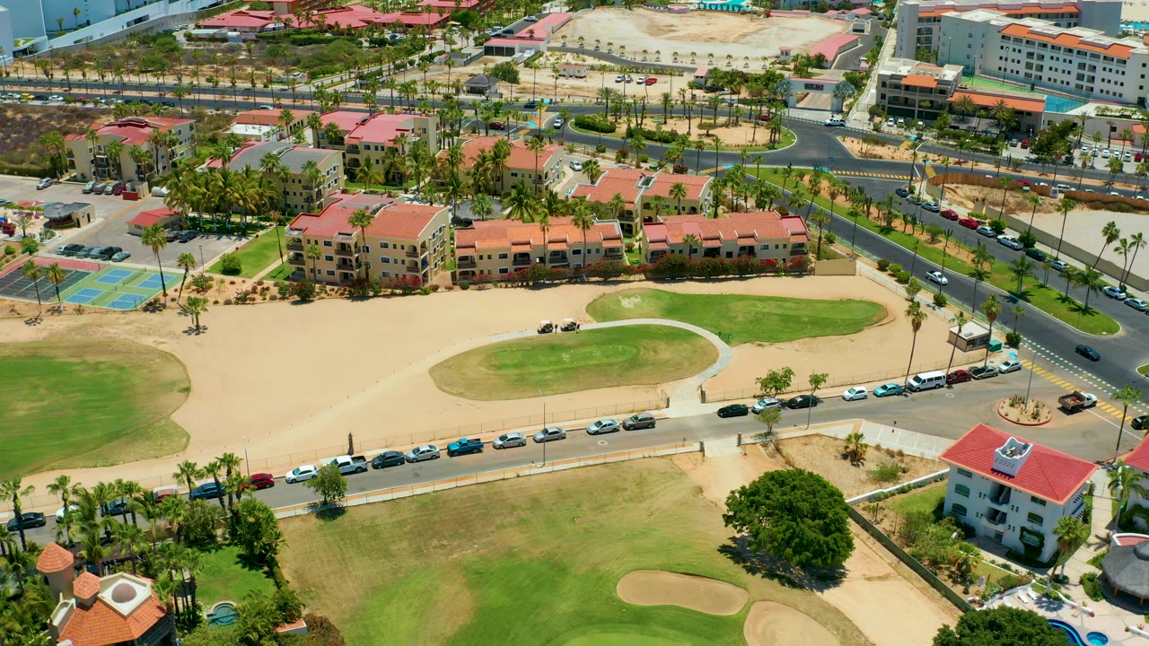 Aerial view in San Jose Del Cabo showing a golf green and sand areas in the foreground, a road with moving and parked cars in the middle, red-roofed buildings behind it, and no visible people.
