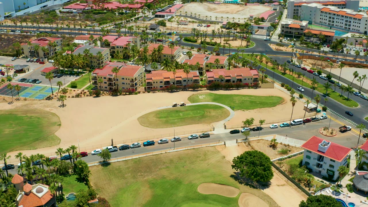 Elevated San Jose Del Cabo frame with a green golf area and sand trap below, a line of parked cars along the road, traffic on the cross street, palm trees, red roofs, and no visible people.