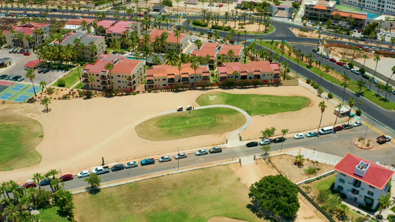 One second into the San Jose Del Cabo aerial, the apartment row and curved street remain centered, with parked cars, a blue court area, palms, green lawns, tan ground, and no visible people.