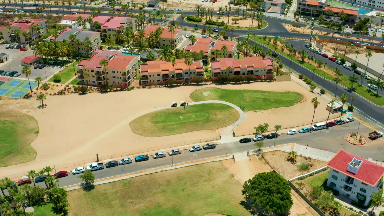 At 0:02, the San Jose Del Cabo frame shows the apartment complex, a nearby road intersection, parked cars along the curb, palm-lined landscaping, green lawn areas, and no visible people.