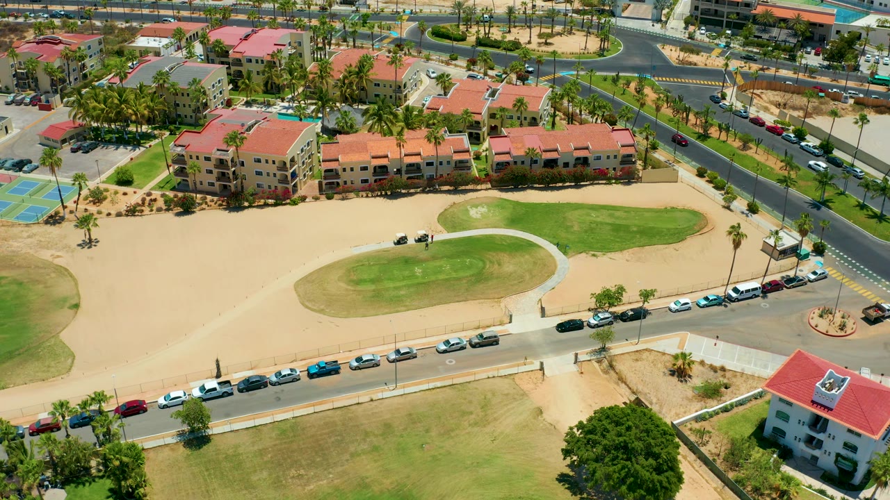 Overhead frame of red-roof buildings, a curved road with cars, palms, green grass, and tan open ground.