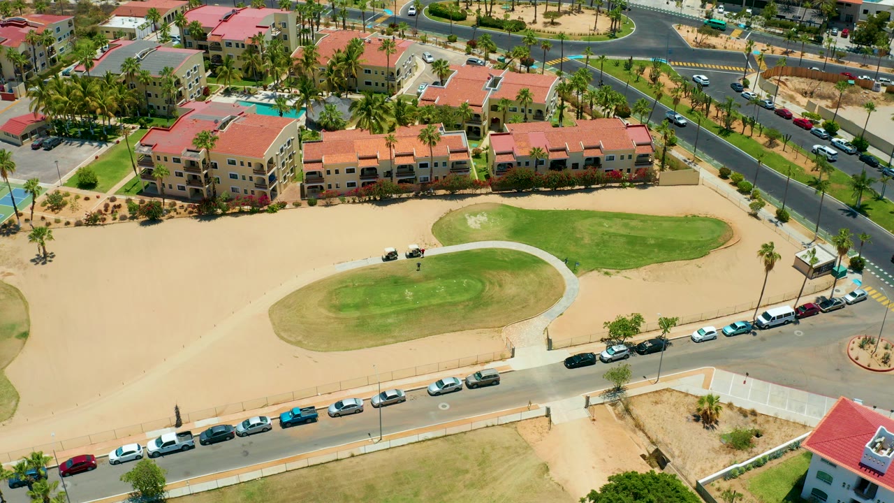 Aerial frame over San Jose Del Cabo showing red-roof residential buildings, palm trees, sandy ground, a green golf area, curved roads with cars, and no visible people.