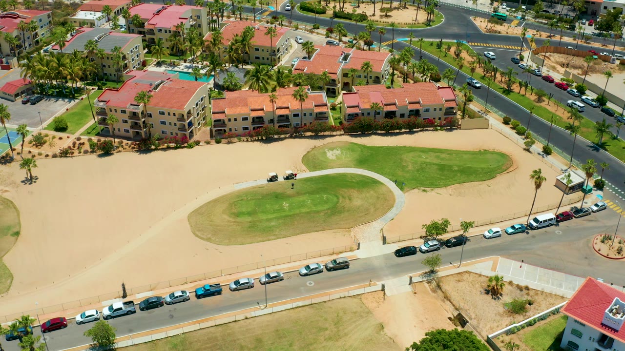 Drone view in San Jose Del Cabo with the oval golf green centered, sandy lots around it, roadside cars along the bottom edge, red-roof homes at the top, and no visible people.