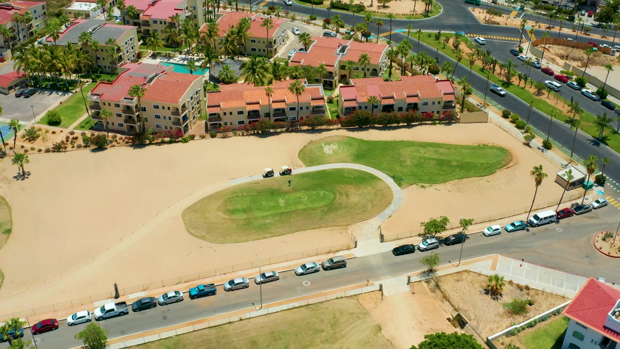 High aerial frame over San Jose Del Cabo showing red-roof residences beyond the golf green, palm-lined streets, parked and moving cars, sandy ground, and no visible people.