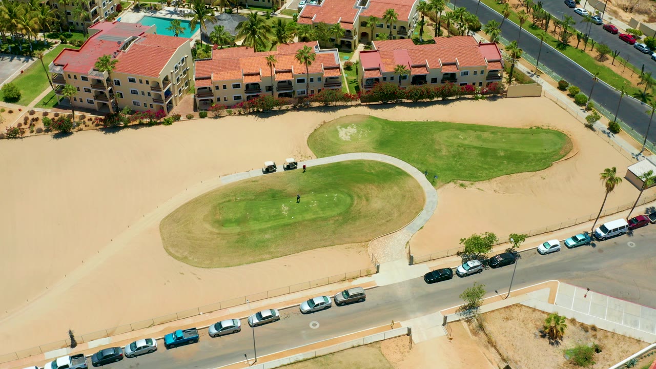 Overhead frame in San Jose Del Cabo showing red-roofed apartment buildings, a sandy courtyard, two green lawn areas, a curved path, parked cars, and no visible people.
