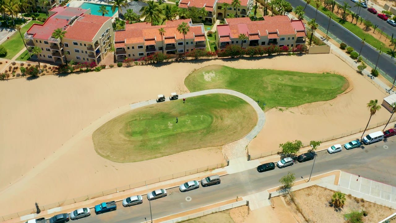 Drone-style overhead frame in San Jose Del Cabo showing red-roofed residential buildings, bright sand, green grass islands, a roadside row of cars, and no visible people.