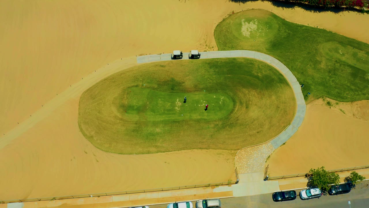 Aerial view of two small people on a green oval field beside sandy ground.