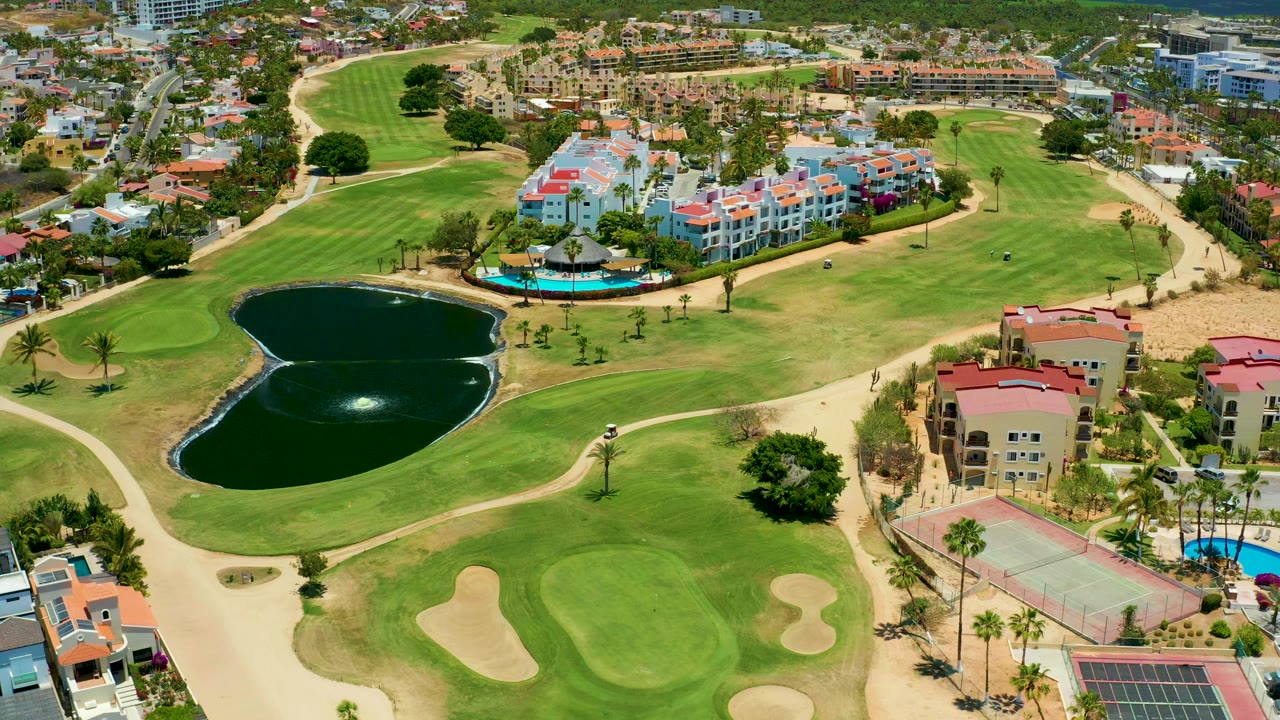 Elevated drone frame in San Jose Del Cabo with the pond slightly left of center, fairways and sand traps below, residential buildings beyond, tennis courts at lower right, and no visible people.