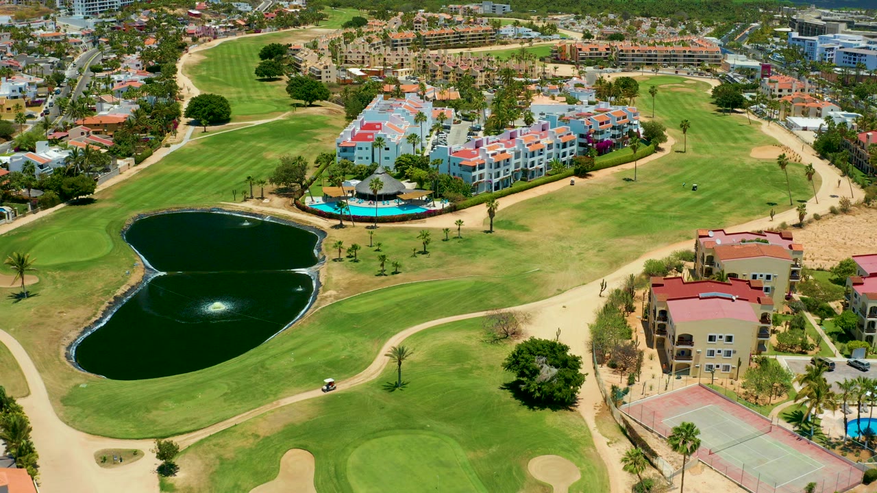 Aerial frame over a San Jose del Cabo golf course with twin dark ponds in the foreground, green fairways, white and red-roofed buildings, a blue pool, and distant residential blocks.