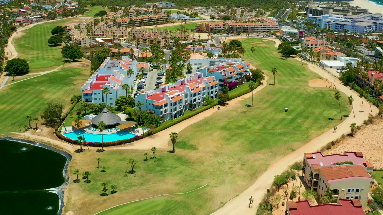 Aerial frame over San Jose del Cabo showing white residential buildings, red roofs, a turquoise pool, green fairway, sandy paths, pond edge, and no visible people.