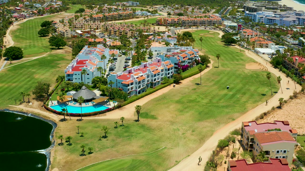 Aerial frame in San Jose del Cabo with the pool and residential buildings slightly more centered, golf-course grass across the foreground, pond edge at left, and no visible people.