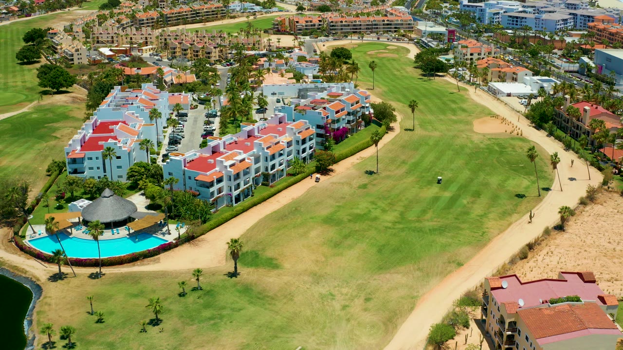 Aerial view in San Jose Del Cabo showing white residential buildings with red roofs, a turquoise pool at lower left, a tan path, and green golf fairways beyond.