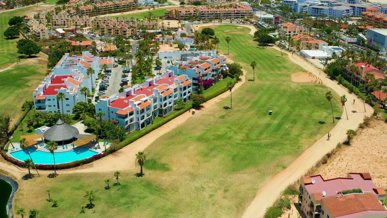 San Jose Del Cabo aerial view with white apartment-style buildings at left, a small pool area below, tan cart paths, sand bunkers, and green fairways stretching to the right.