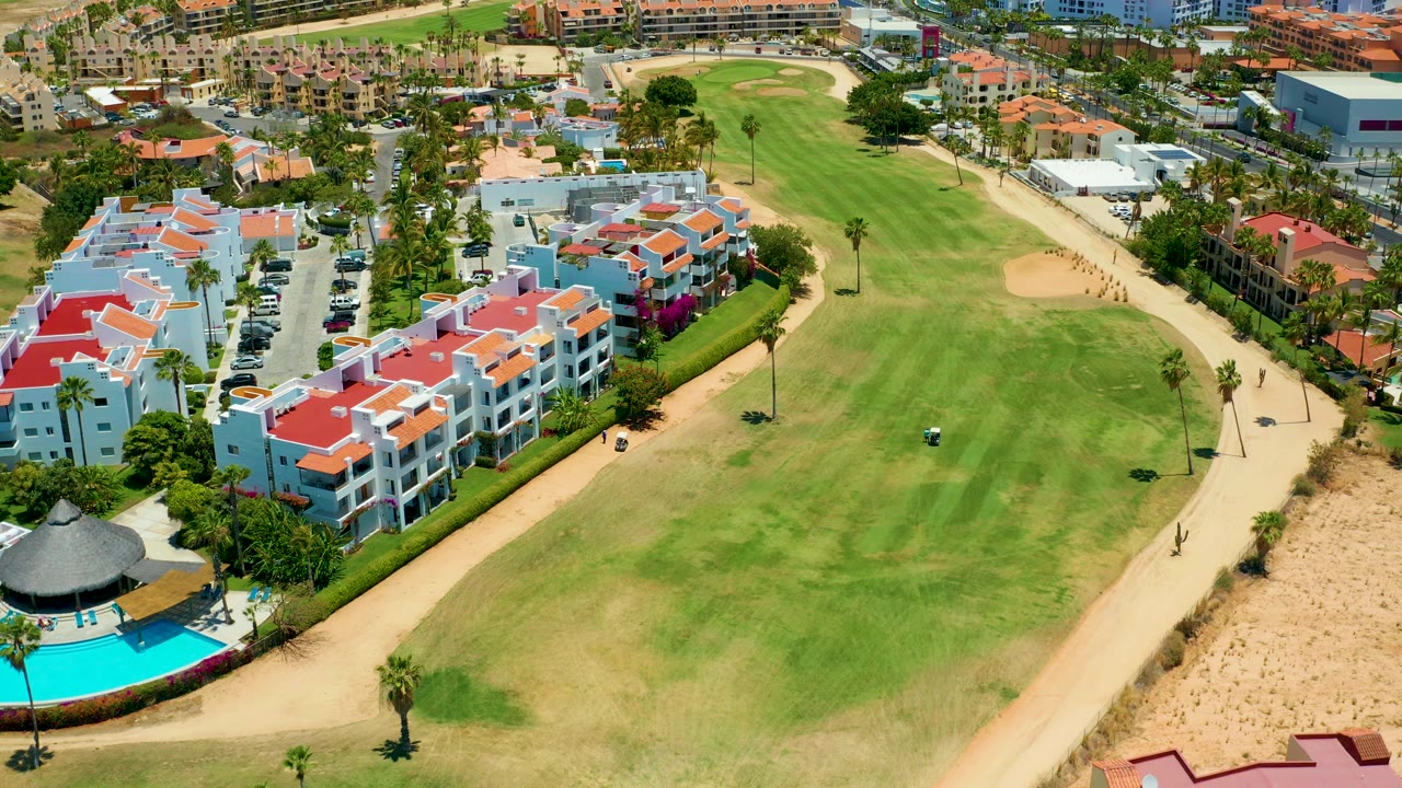 San Jose Del Cabo aerial still with the fairway centered, blue and white residential villas on the left, tan paths along the green, palm trees, parked cars, and distant urban buildings.