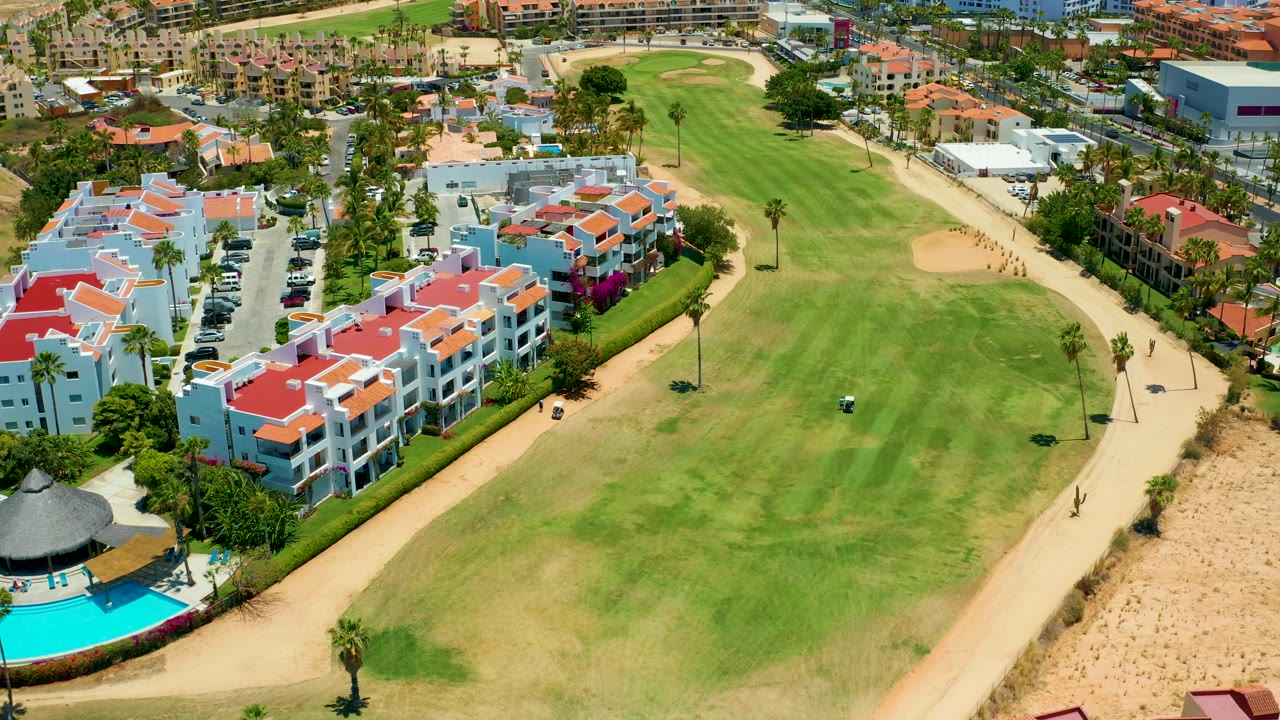 Aerial San Jose Del Cabo frame with a broad green fairway in the center, red-roofed homes to the left and upper right, sandy ground along the right edge, and a small dark golf cart on the grass.