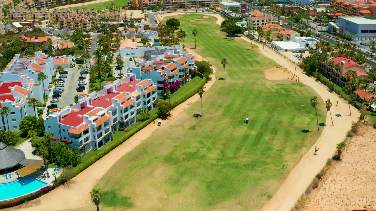 Golf course bordered by apartments, pool area, palm trees, and sandy path.