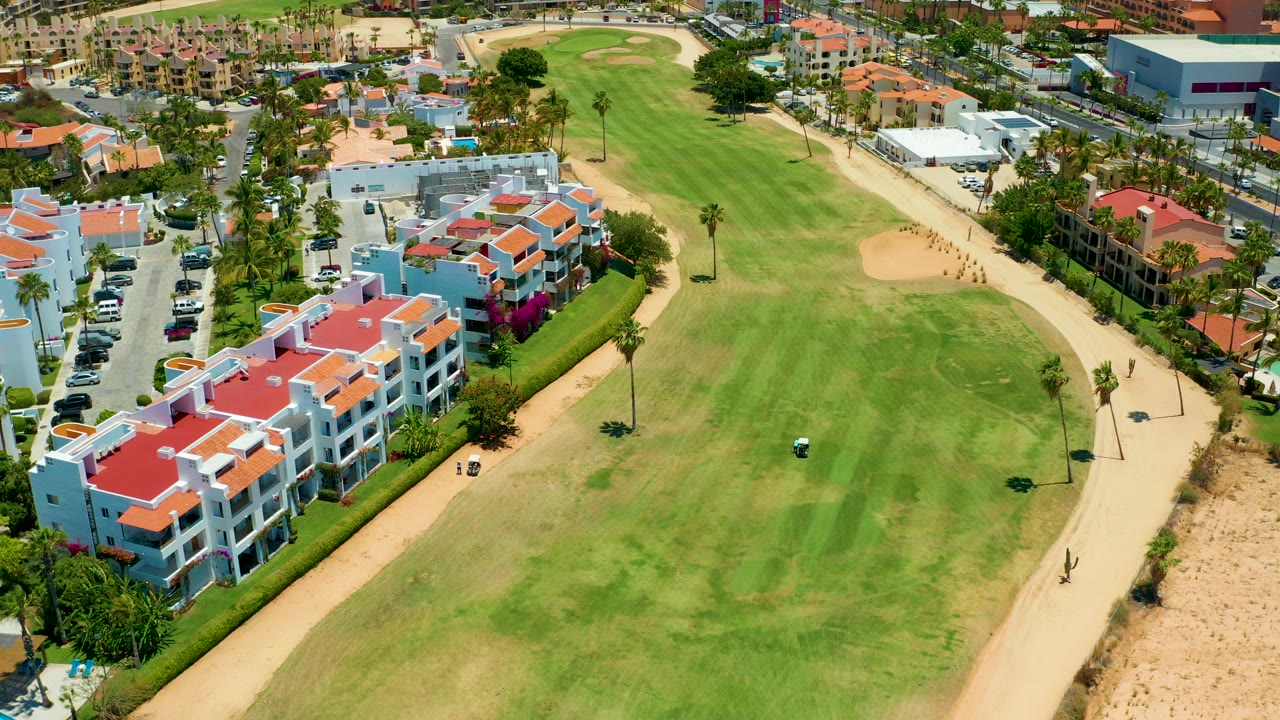 Aerial view in San Jose Del Cabo showing a green golf fairway, sandy cart path, red-roofed white buildings, palm trees, parked vehicles, and no visible people.